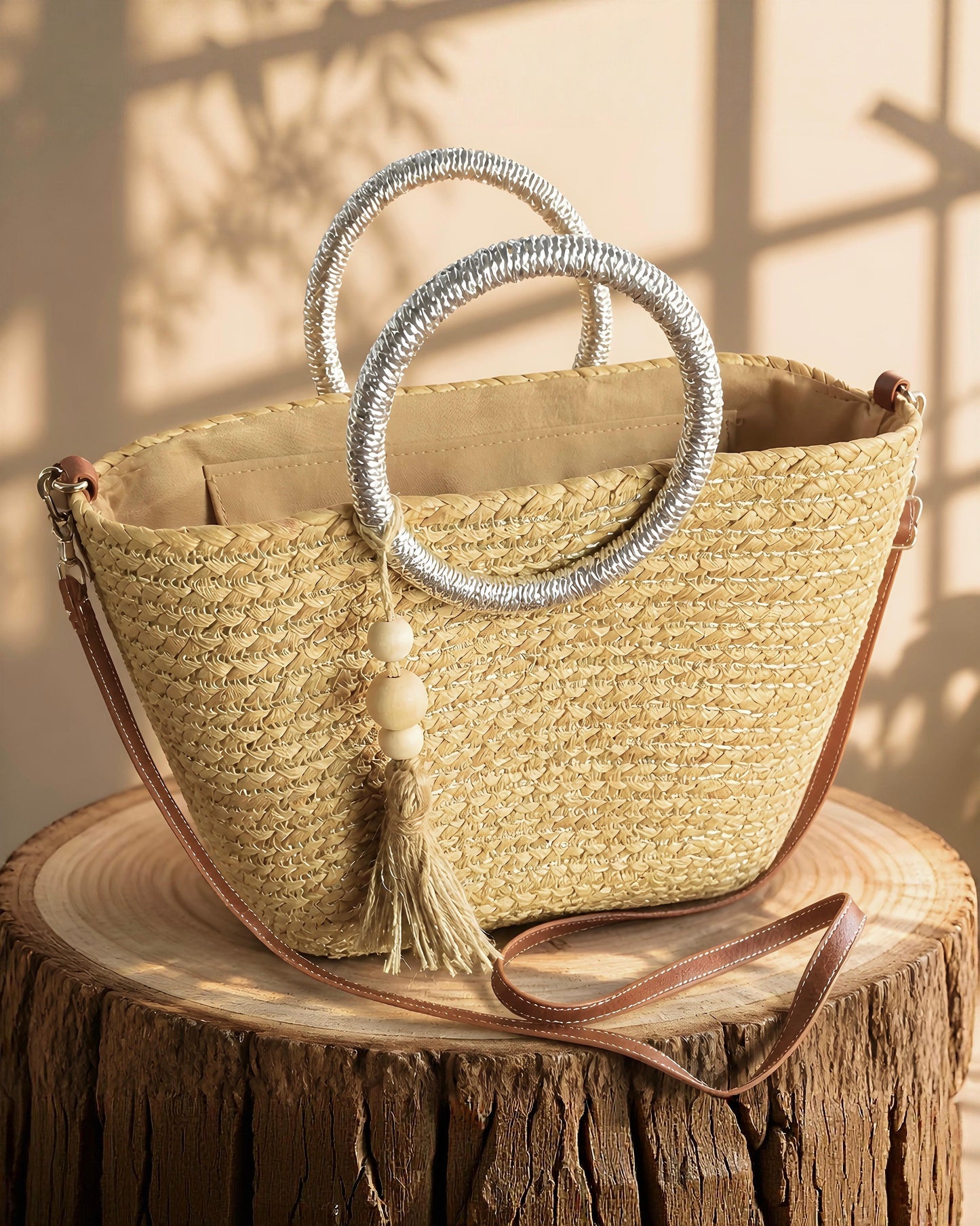 Bolso capazo de paja modelo Pallejá de la marca EUTEA con un cuerpo trenzado natural, llamativas asas circulares de color plata brillante y una borla de hilo con cuentas de madera, expuesto en un ambiente mediterráneo con arena fina y caracolas.