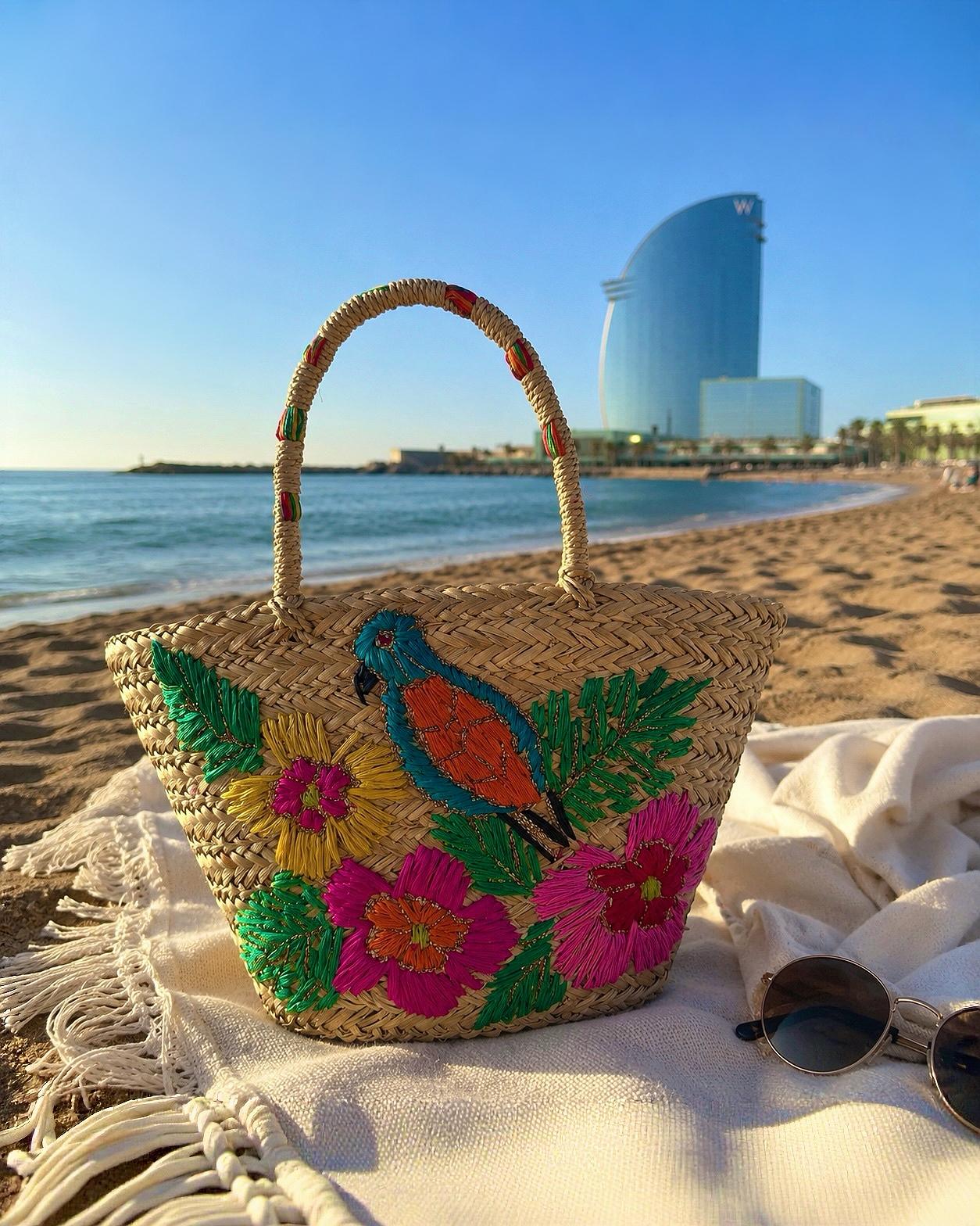 Bolso estilo capazo de paja modelo San Pol de la marca EUTEA con un colorido bordado de un pájaro azul y flores tropicales amarillas y rosas, fotografiado en la arena de la playa de Barcelona.
