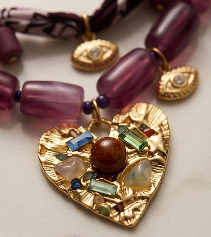 Primer plano del Collar Pals con colgante dorado en forma de corazón y piedras decorativas sobre tejido satinado.