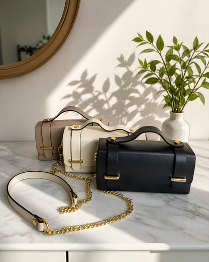 Bolso bandolera estructurado de cuero modelo Cunit de la marca EUTEA, expuesto en tres colores (negro, blanco marfil y beige camel) sobre una superficie de mármol con elegantes herrajes dorados y correa de cadena combinada.