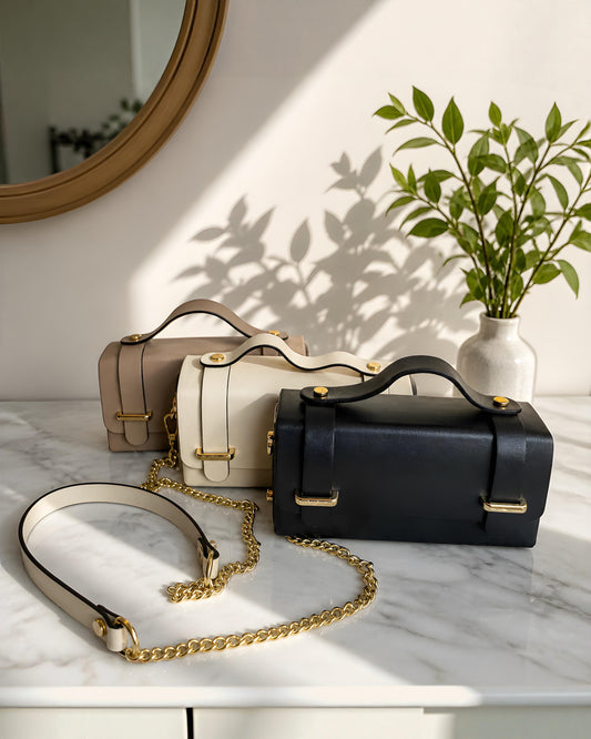 Bolso bandolera estructurado de cuero modelo Cunit de la marca EUTEA, expuesto en tres colores (negro, blanco marfil y beige camel) sobre una superficie de mármol con elegantes herrajes dorados y correa de cadena combinada.