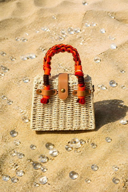 Bolso de mano de mimbre natural Taormina expuesto sobre la arena de la playa con esferas de cristal decorativas.