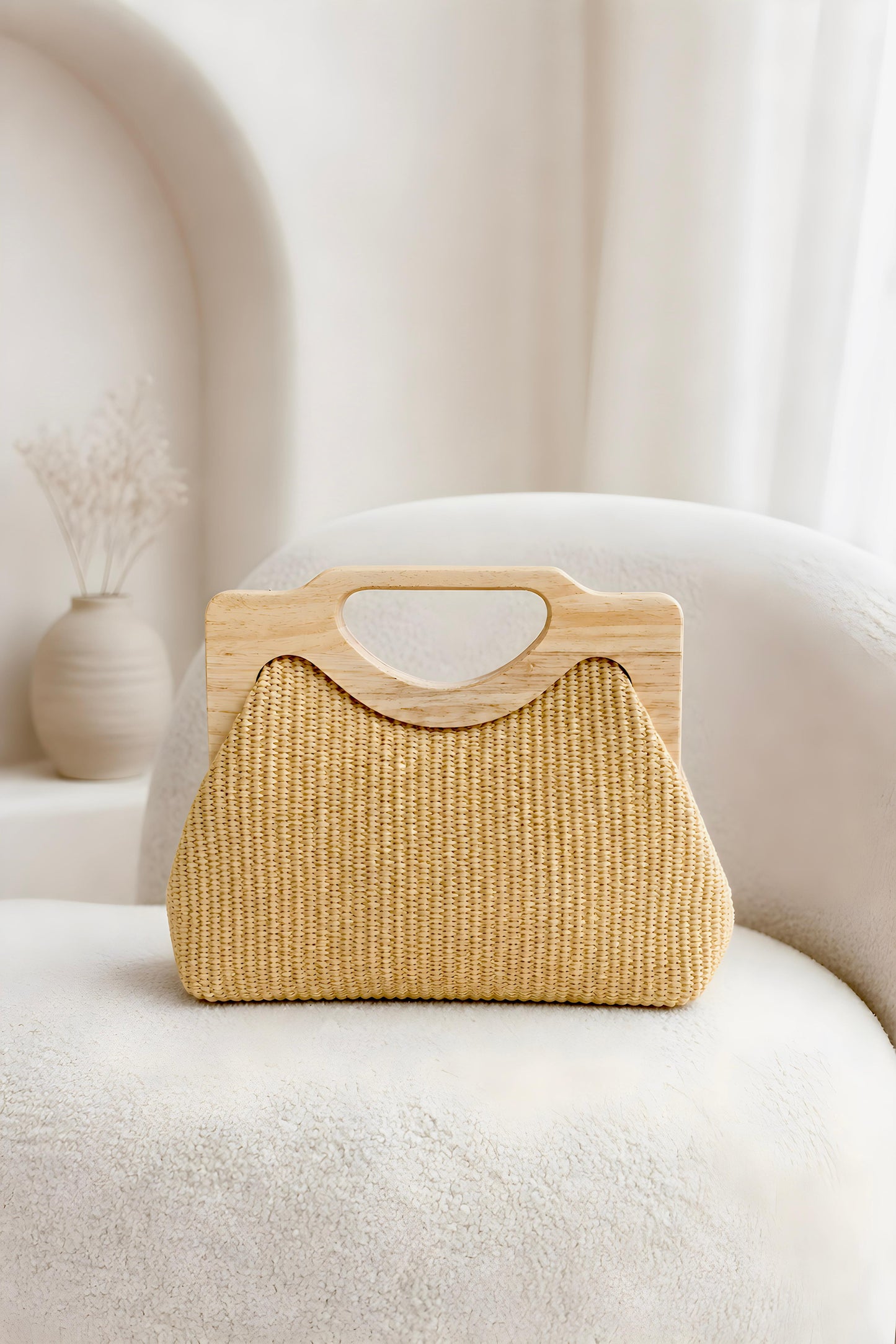 Vista frontal del bolso de mano de rafia natural Positano con asa geométrica de madera clara sobre un sillón blanco.