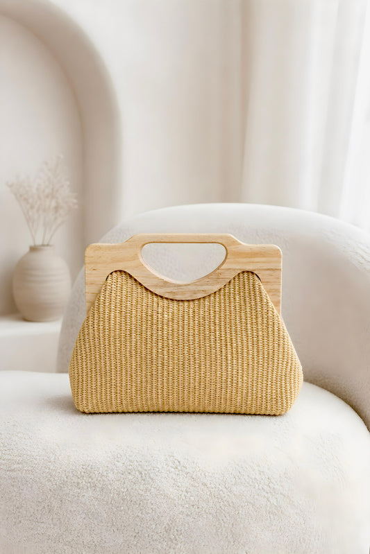 Vista frontal del bolso de mano de rafia natural Positano con asa geométrica de madera clara sobre un sillón blanco.