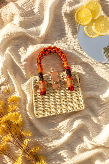 Vista superior del bolso de mimbre rígido Taormina con asa de cordón trenzado multicolor y rodajas de limón sobre fondo textil