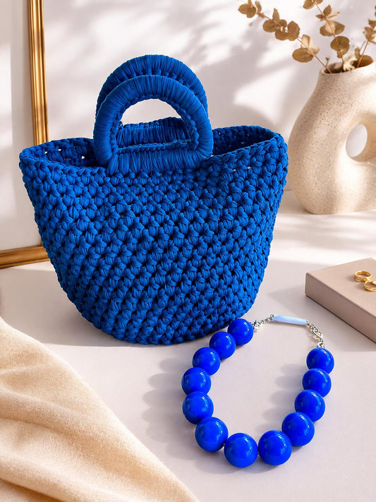 Bolso de mano estilo tote modelo Balis de la marca EUTEA, confeccionado en punto trenzado grueso de color azul eléctrico 100% poliéster, con dos asas de mano superiores y expuesto junto a un collar de grandes esferas azules a juego.