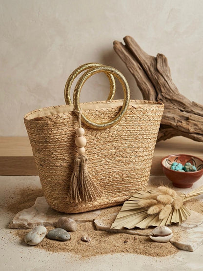 Bolso capazo de paja modelo Pallejá de la marca EUTEA con un cuerpo trenzado natural, llamativas asas circulares de color oro brillante y una borla de hilo con cuentas de madera, expuesto en un ambiente mediterráneo con arena fina y caracolas.