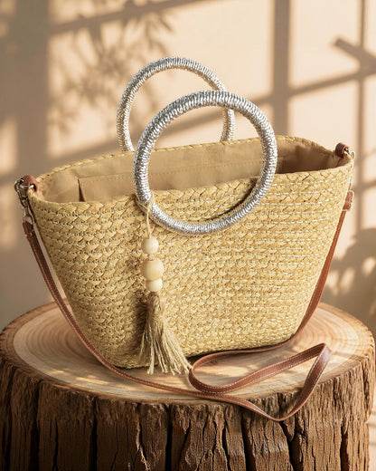 Bolso capazo de paja modelo Pallejá de la marca EUTEA con un cuerpo trenzado natural, llamativas asas circulares de color plata brillante y una borla de hilo con cuentas de madera, expuesto en un ambiente mediterráneo con arena fina y caracolas.