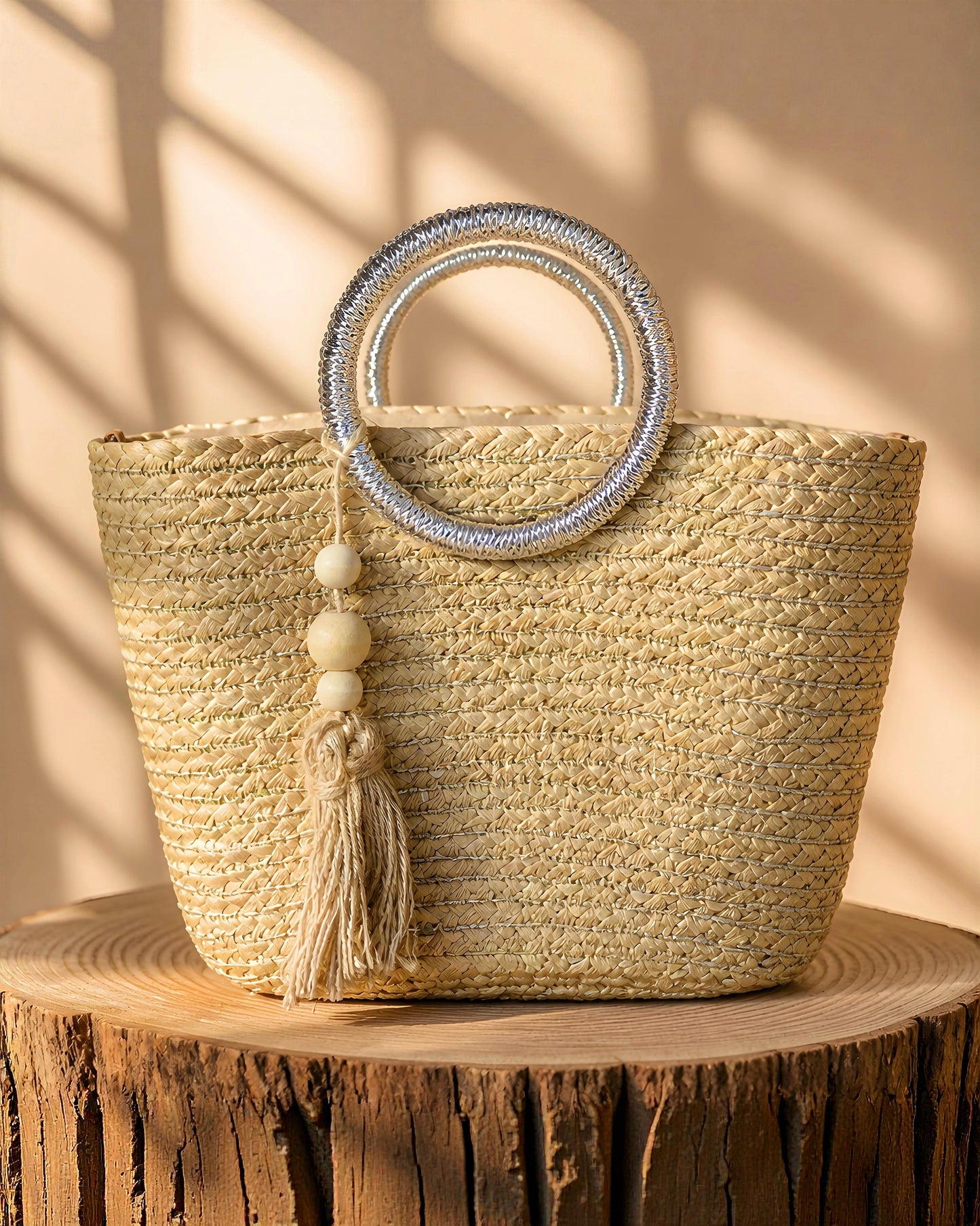 Bolso capazo de paja modelo Pallejá de la marca EUTEA con un cuerpo trenzado natural, llamativas asas circulares de color plata brillante y una borla de hilo con cuentas de madera, expuesto en un ambiente mediterráneo con arena fina y caracolas.