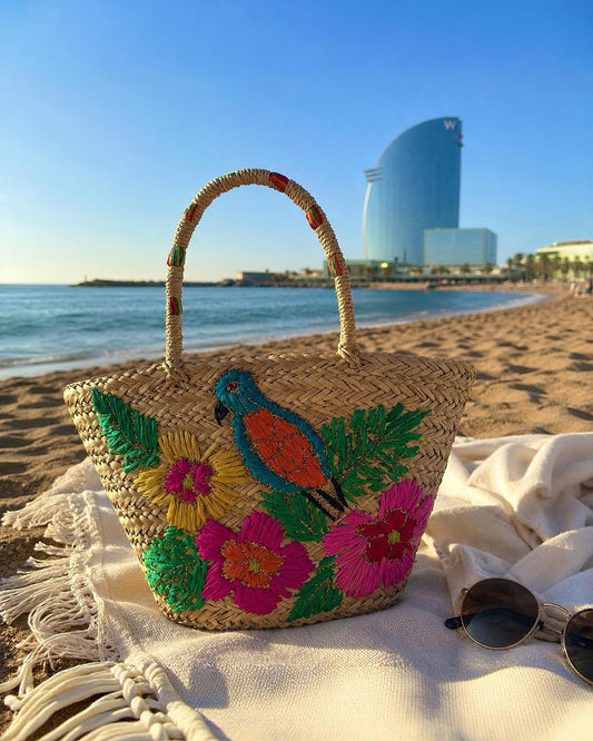 Bolso estilo capazo de paja modelo San Pol de la marca EUTEA con un colorido bordado de un pájaro azul y flores tropicales amarillas y rosas, fotografiado en la arena de la playa de Barcelona.