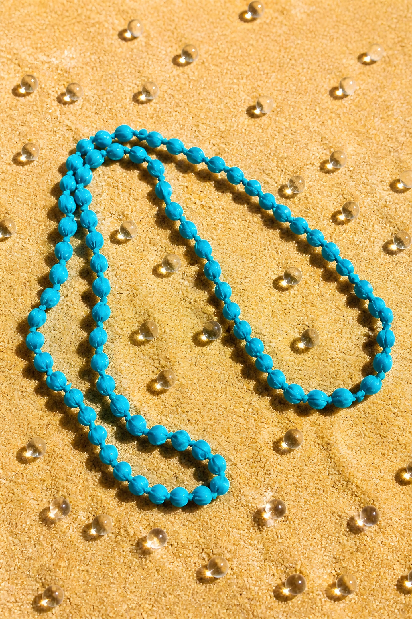 Collar largo de paño y madera Begur en color azul turquesa extendido sobre un fondo de arena con esferas transparentes.