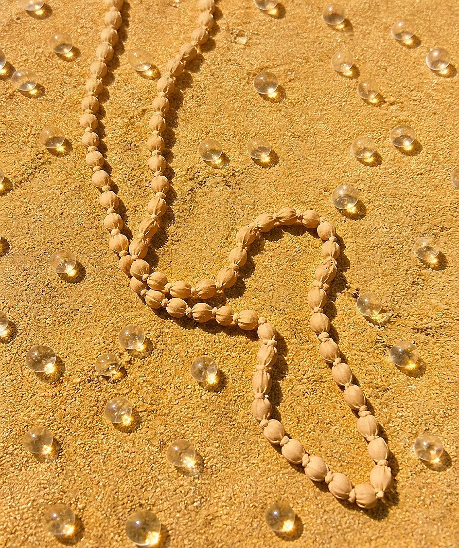 Collar artesanal largo Begur en tono beige arena, elaborado con cuentas de madera y tela suave sobre superficie de arena.