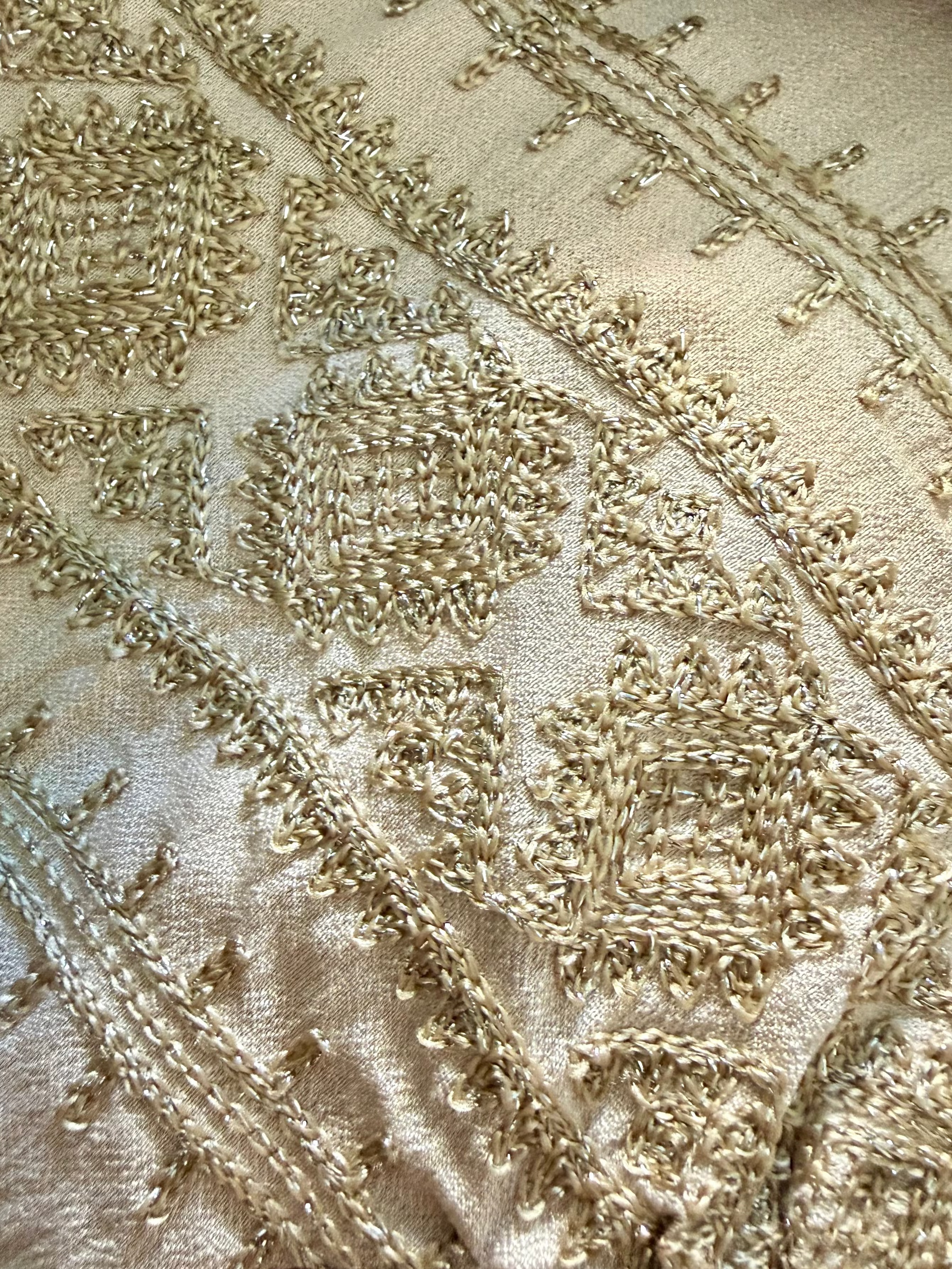 Vestido corto a la rodilla Alella en color beige de la marca EUTEA, mostrando el escote cruzado en V, el lazo de la cintura y los bordados en tonos beige y oro, posando descalza frente a una pared blanca soleada