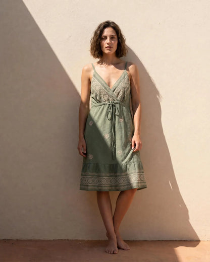 Vestido corto a la rodilla Alella en color verde caqui de la marca EUTEA, mostrando el escote cruzado en V, el lazo de la cintura y los bordados en tonos beige y oro, posando descalza frente a una pared blanca soleada