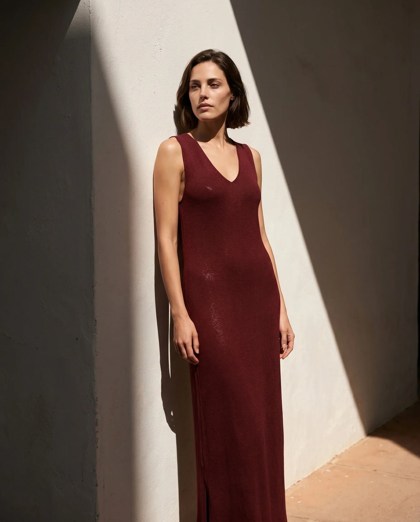 Vestido maxi de punto fino Corrals en color granate de la marca EUTEA, de silueta recta sin mangas y escote en V, apoyado en una pared blanca con sombras marcadas por el sol