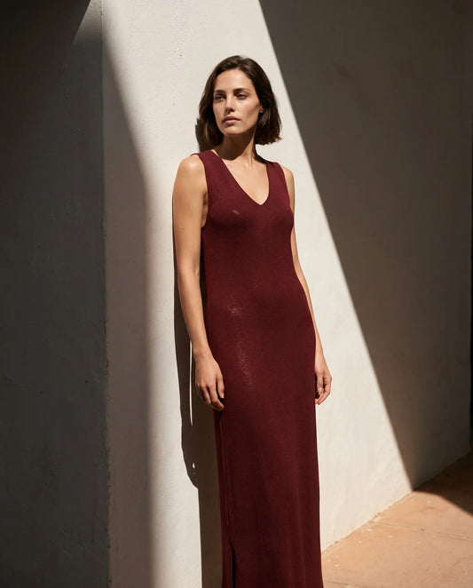 Vestido maxi de punto fino Corrals en color granate de la marca EUTEA, de silueta recta sin mangas y escote en V, apoyado en una pared blanca con sombras marcadas por el sol