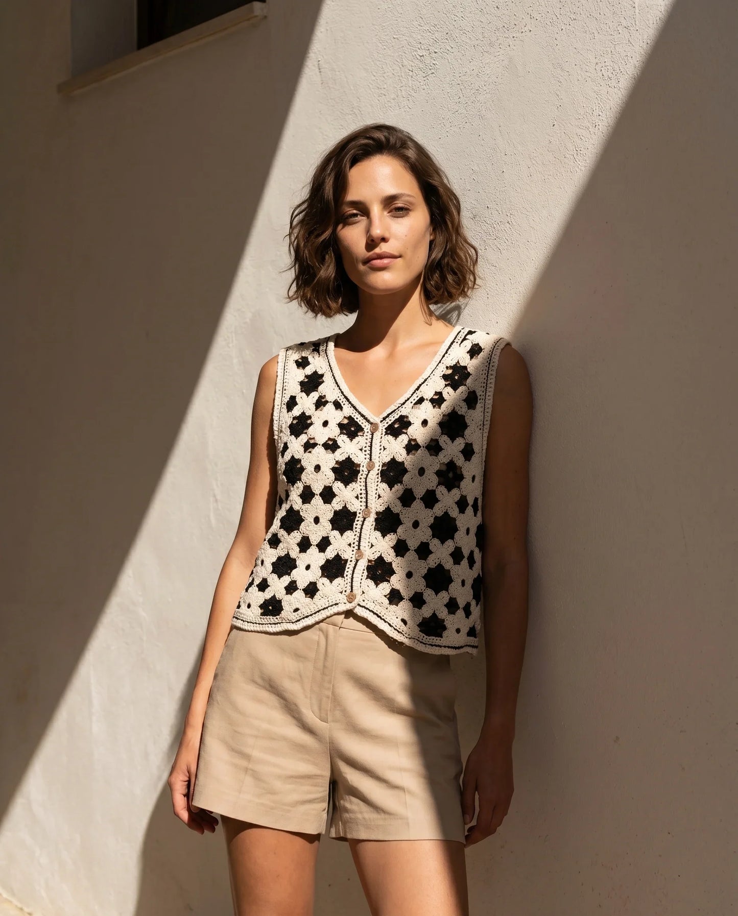 Chaleco calado de crochet Tabarca en contraste crudo y negro de la marca EUTEA, mostrando el escote en V con botones frontales, combinado con unos shorts beige y apoyada en una pared blanca iluminada por el sol