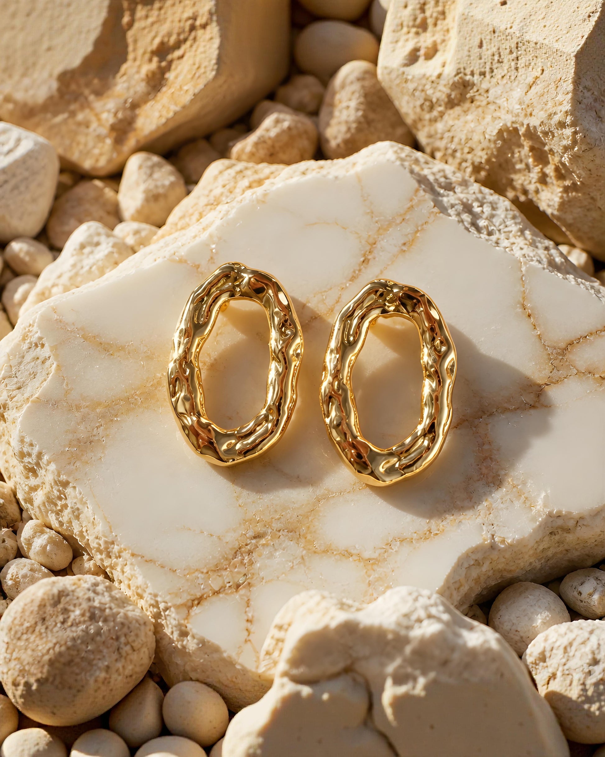 Pendientes ovalados dorados Cadaqués de la marca EuTea, fabricados en acero inoxidable con relieve irregular de metal fundido sobre piedras claras.
